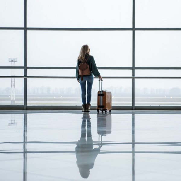 A woman wearing jeans, a jacket and boots while carrying a knapsack and holding a hardshell luggage while looking out the airport window.