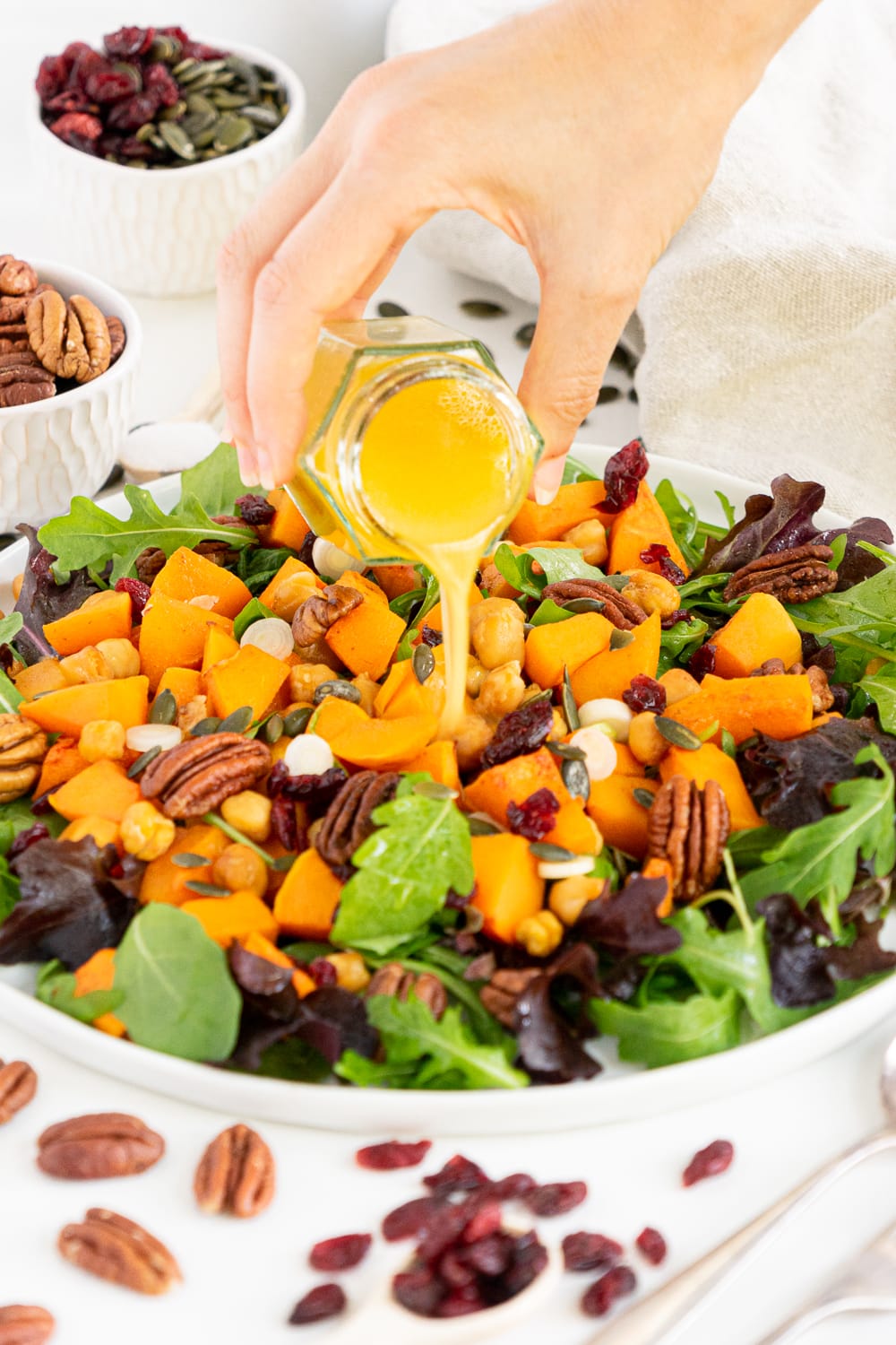 3/4 view of roasted butternut squash salad with arugula, pecans and dried cranberries with hand pouring dressing on it