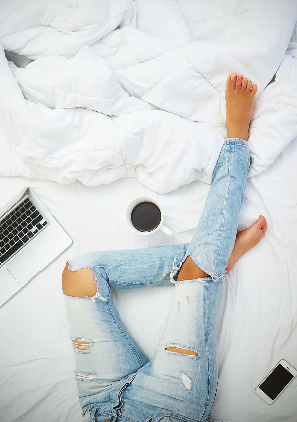 Top view of woman's leg on a bed with a laptop and coffe next to her.