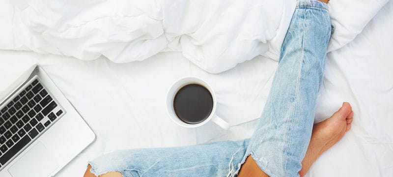 Top view of woman's leg on a bed with a laptop and coffe next to her.