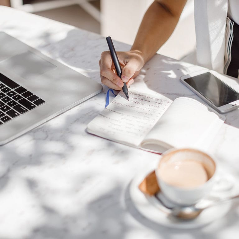 Freelance writer woman writing on a notepad with a laptop next to her.