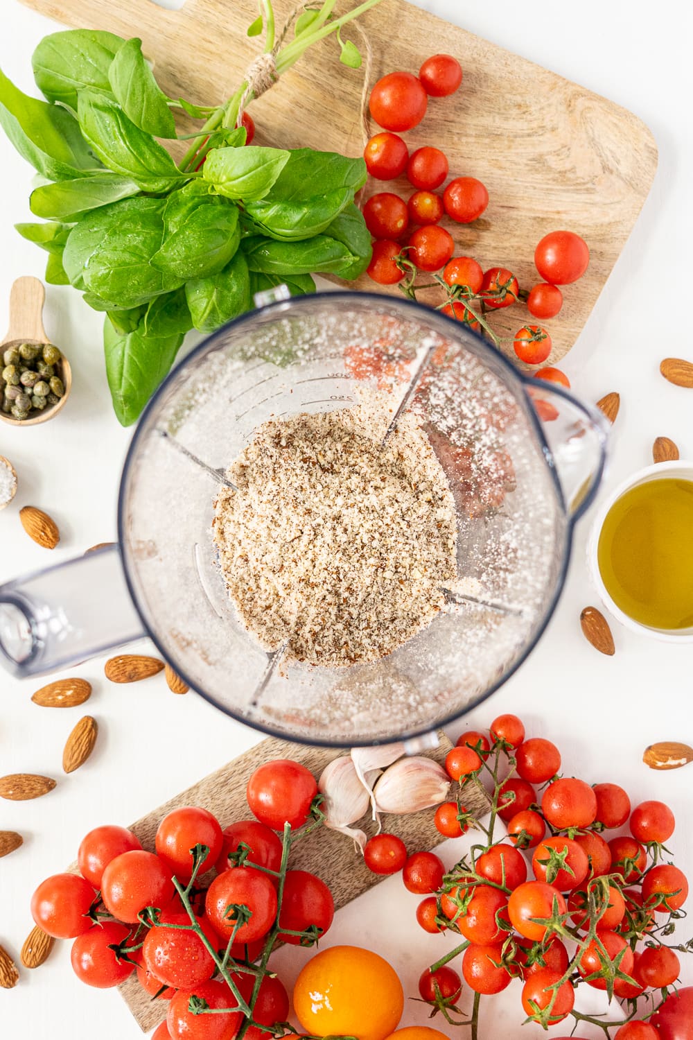 Top view of food processor with grounded almonds inside, surrounded by tomatoes and basil