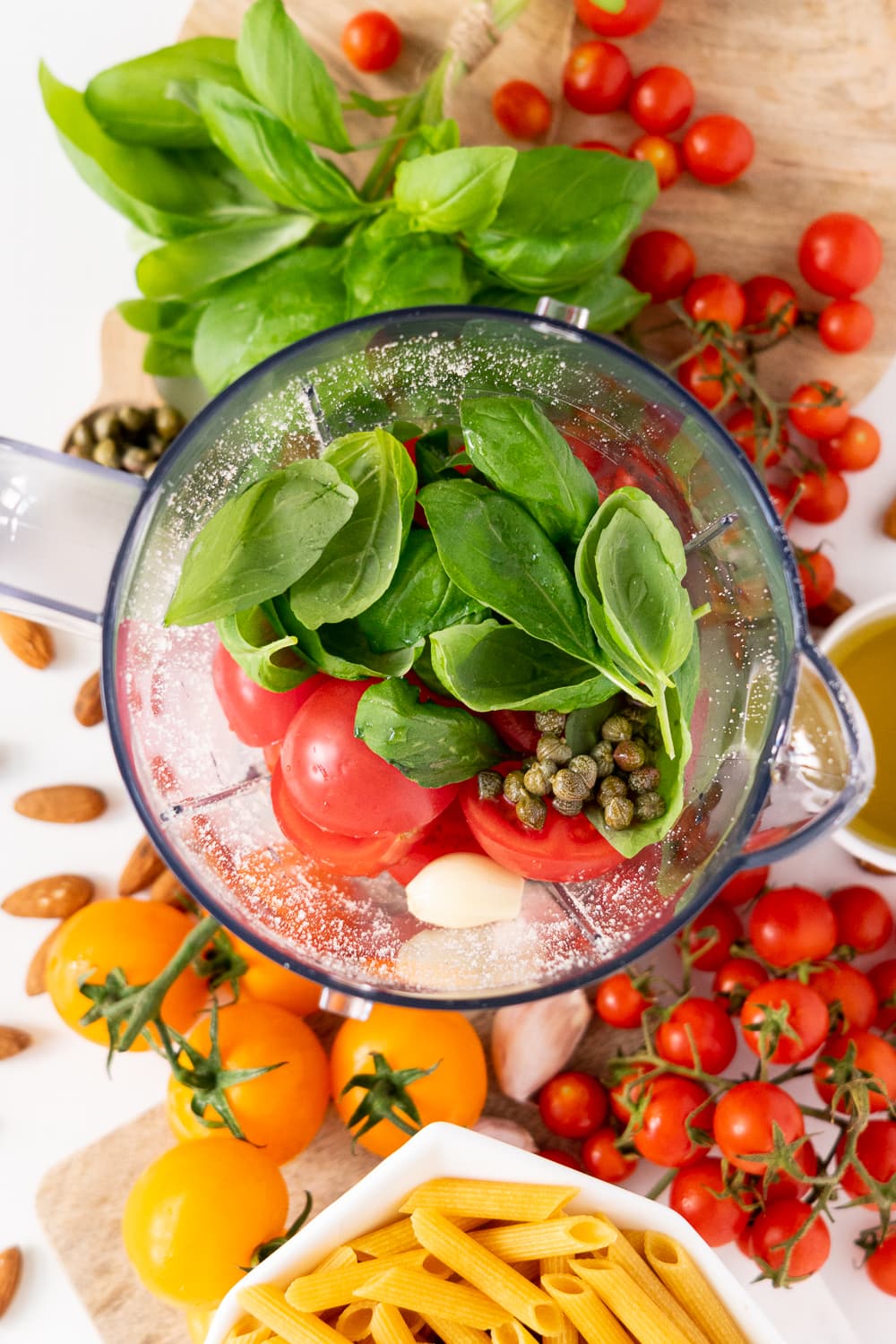 Top view of blender with basil, tomatoes, garlic, capers, olive oil and almonds inside, surrounded by tomatoes and basil