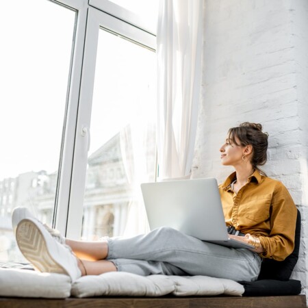 A woman with a laptop looking outside a window while looking for online bookkeeping jobs
