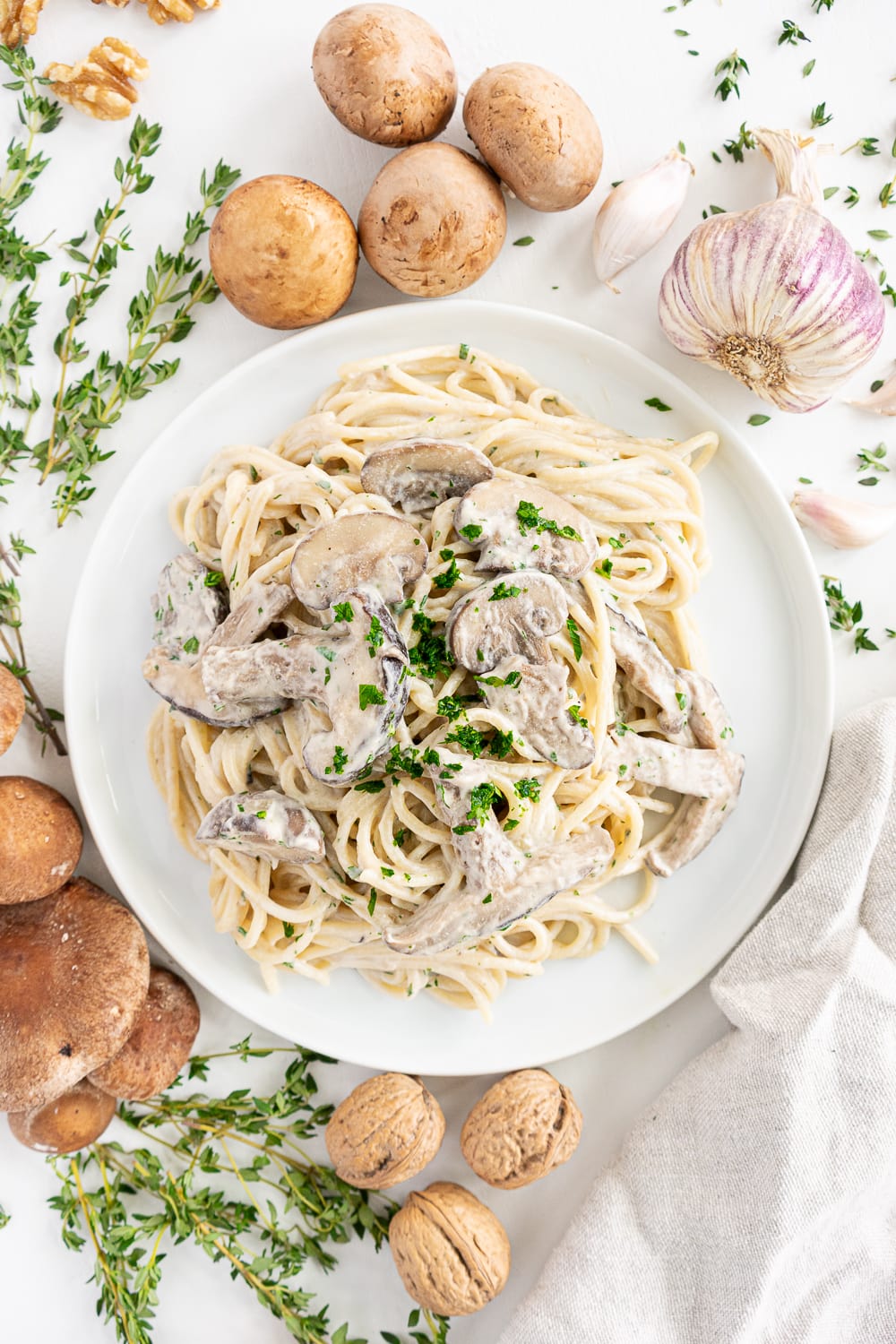 Top view of a white plate with creamy vegan mushroom pasta surrounded by mushrooms, garlic, and thyme