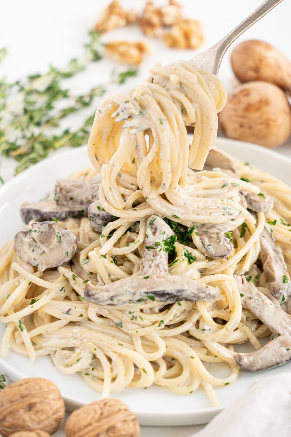 Three quarter view of a white plate with creamy vegan mushroom pasta surrounded by mushrooms, garlic, and thyme with a fork full of pasta