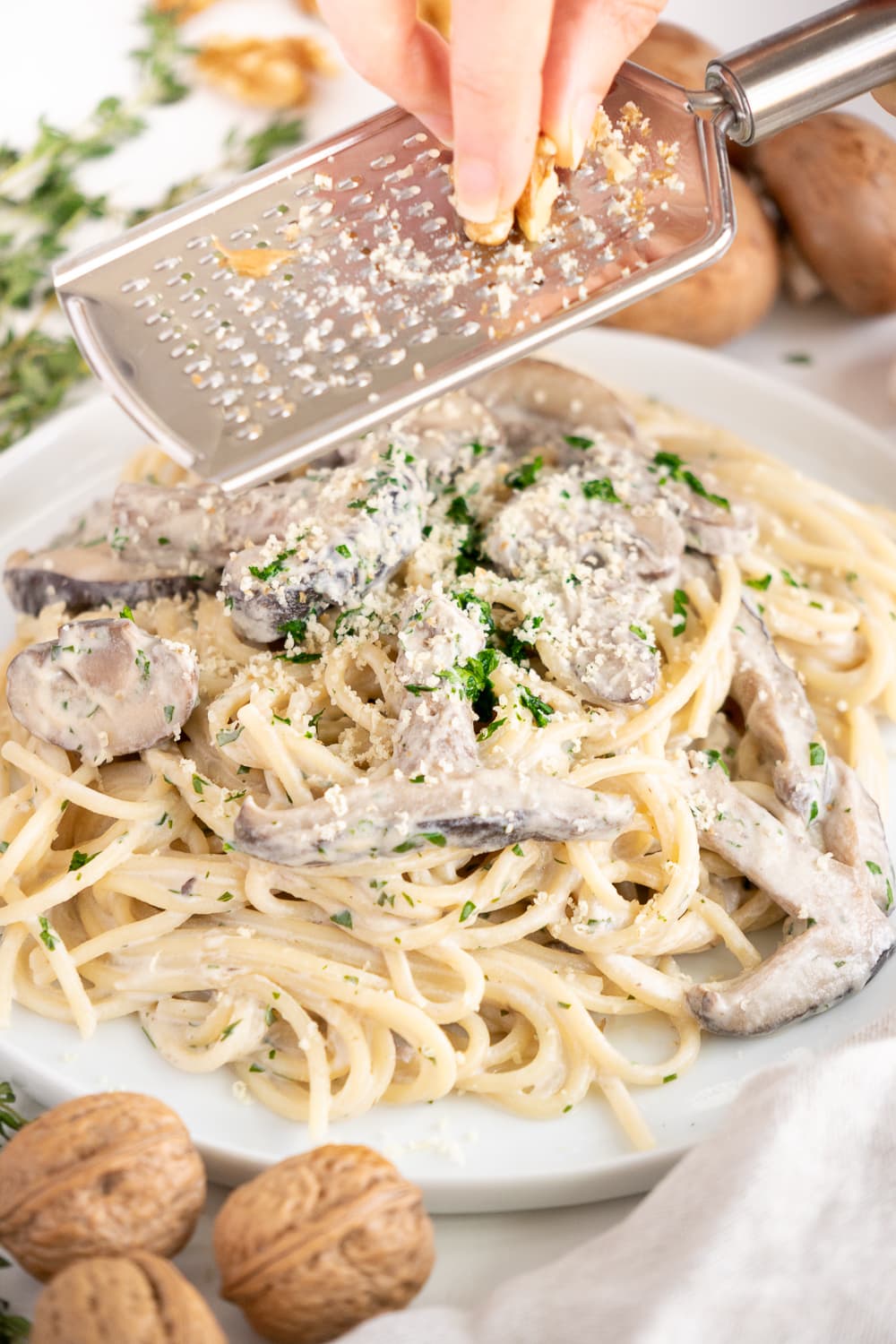 Three quarter view of a white plate with creamy vegan mushroom pasta with hand grating walnuts on top