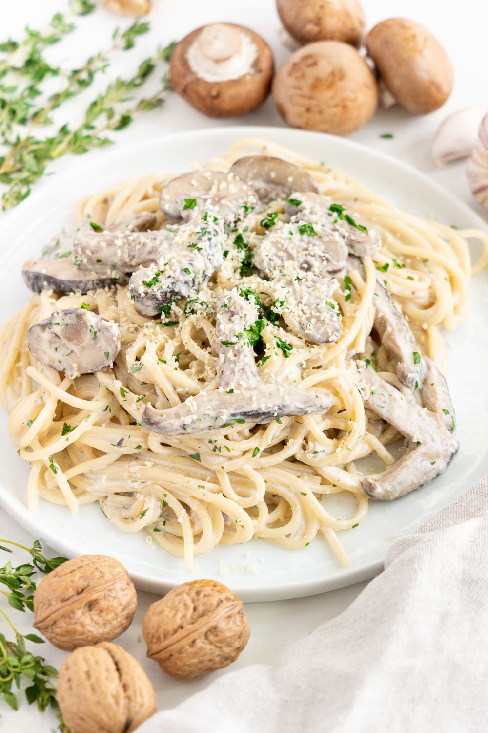 Three quarter view of a white plate with creamy vegan mushroom pasta surrounded by mushrooms, garlic, and thyme