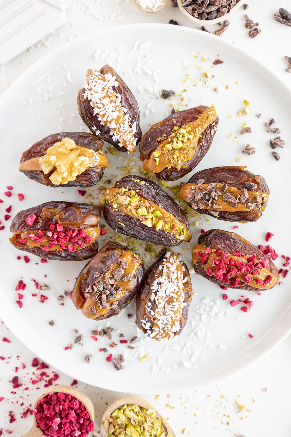 Top view of a white plate with stuffed dates filled with peanut butter, pistachios, coconut flakes, freeze-dried raspberries, and cacao nibs