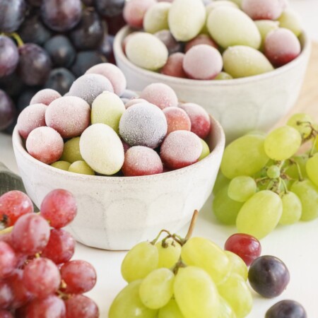 Frozen red and green grapes in white bowls surrounded by fresh red and white grapes.