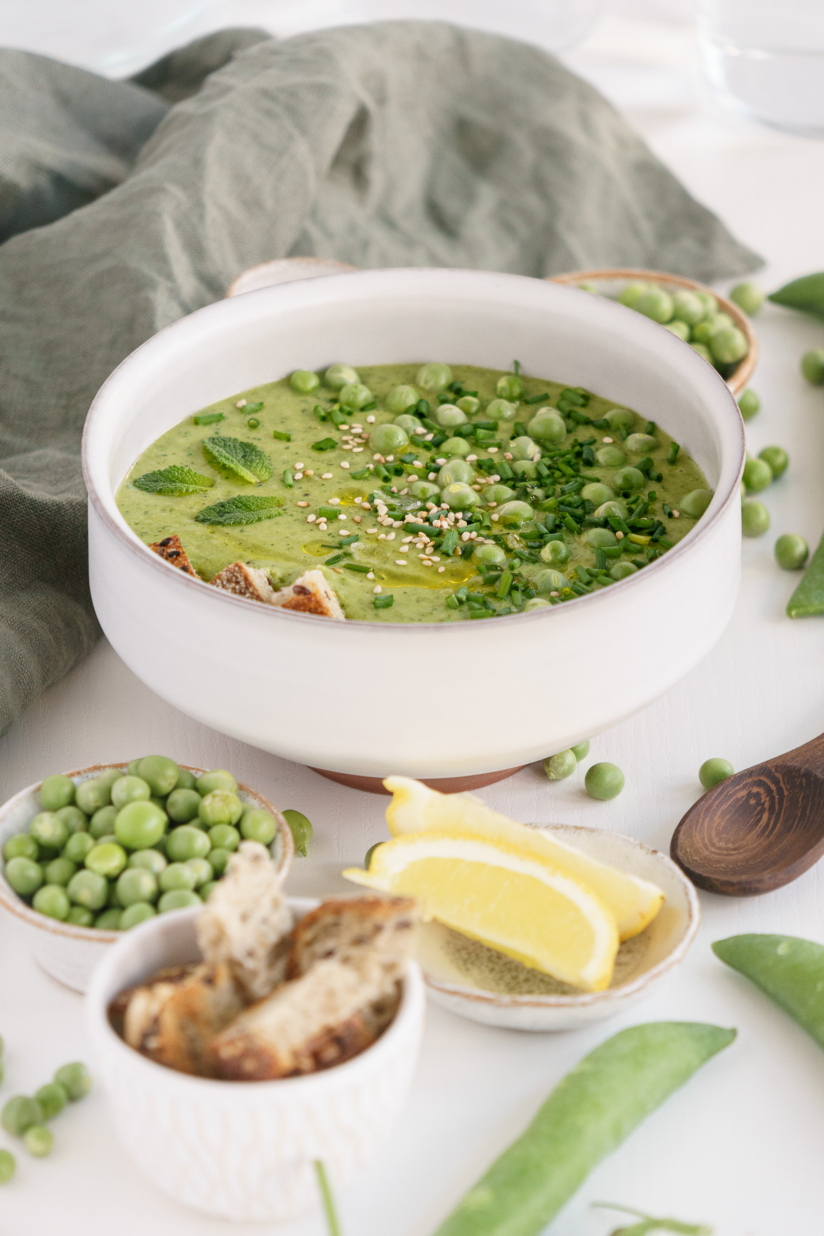 Bowl of fresh pea soup on the table with lemon wedges, croutons and a wooden spoon.