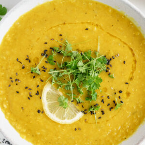 Closeup overhead of bowl of red lentil soup with lemon wedge and spices for garnish.