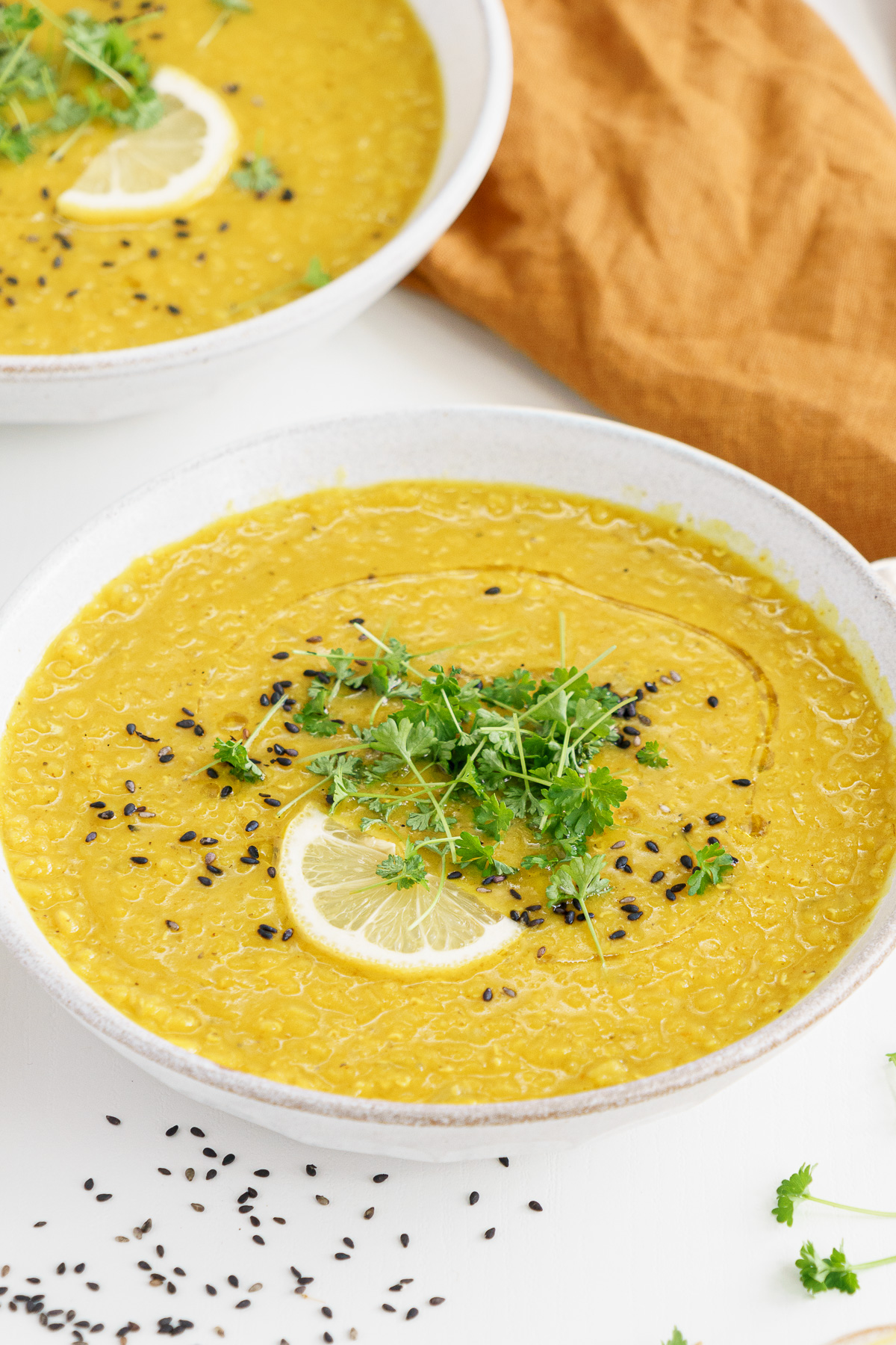 Bowl of middle eastern red lentil soup on the table garnished with slice of lemon and fresh herbs.