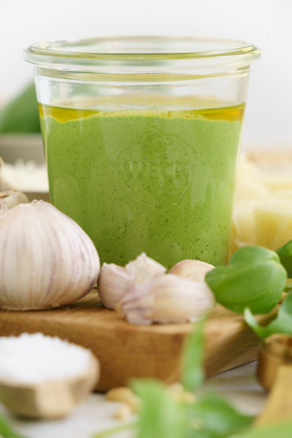 Pesto in a jar on a cutting board with a garlic, basil leaves and cheese around it.