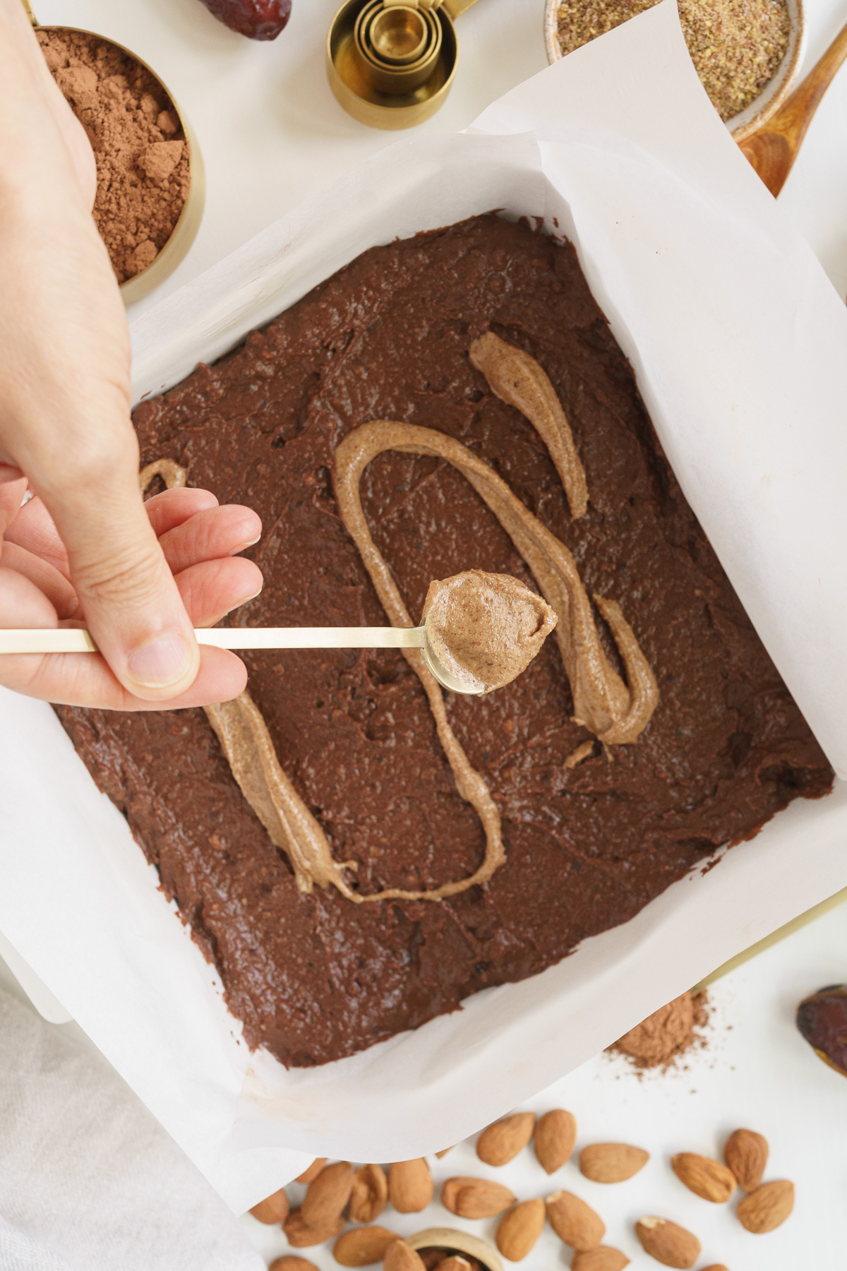 Top view of hand spreading almond butter on top of brownies batter with a spoon