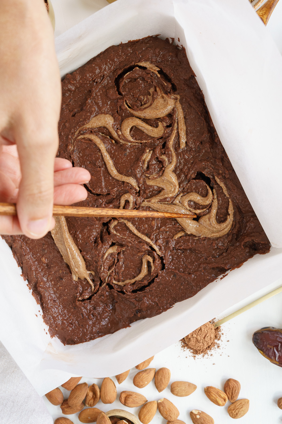 Top view of hand spreading almond butter on top of brownies batter with a chopstick