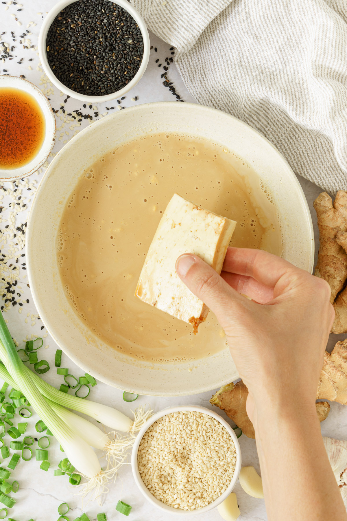 top view of a hand dipping the sliced tofu in the marinade with black and white sesame seeds, rice vinegar, green onions and ginger