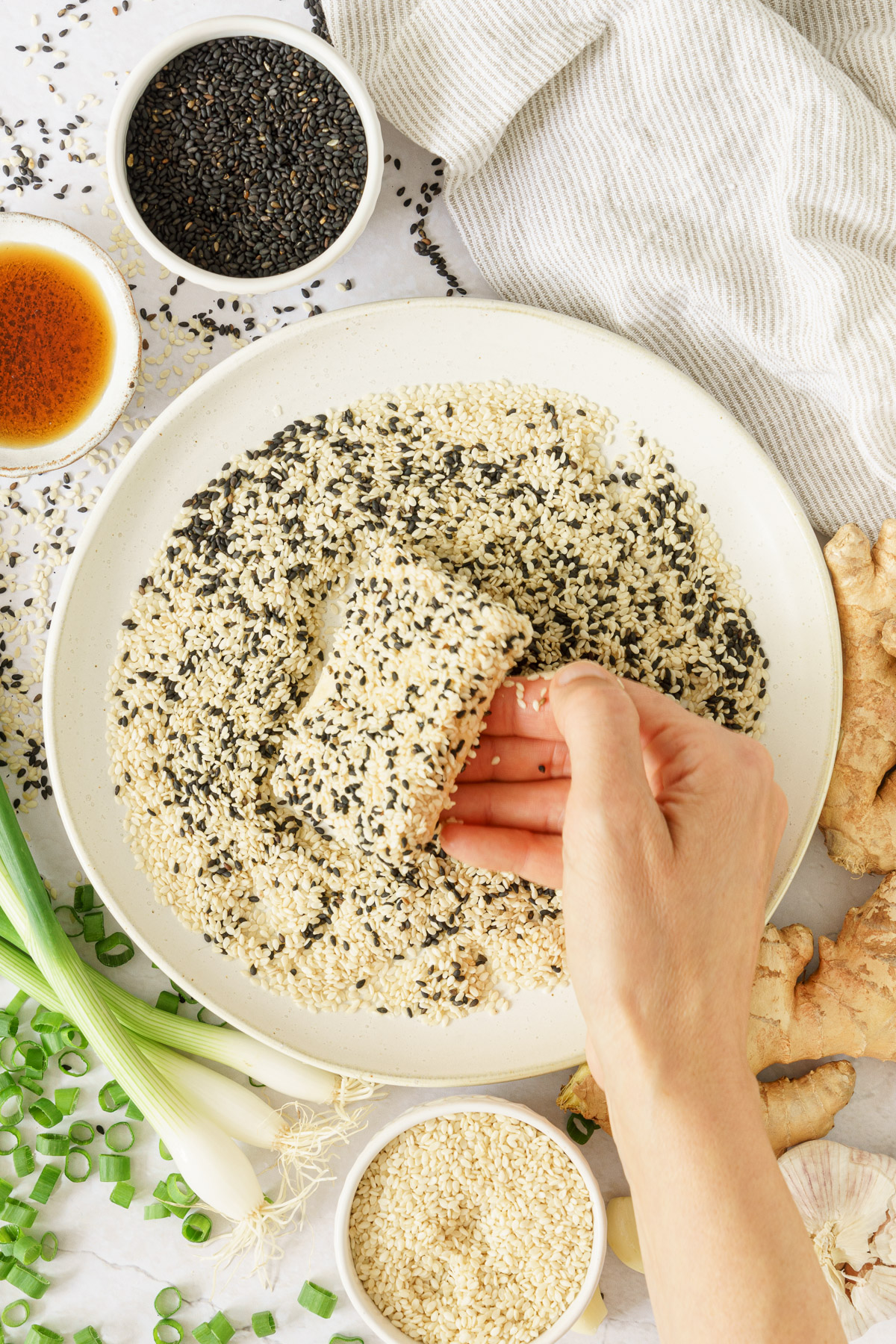 top view of a hand coating the sliced tofu in the black and white sesame seeds mixture with rice vinegar, green onions and ginger