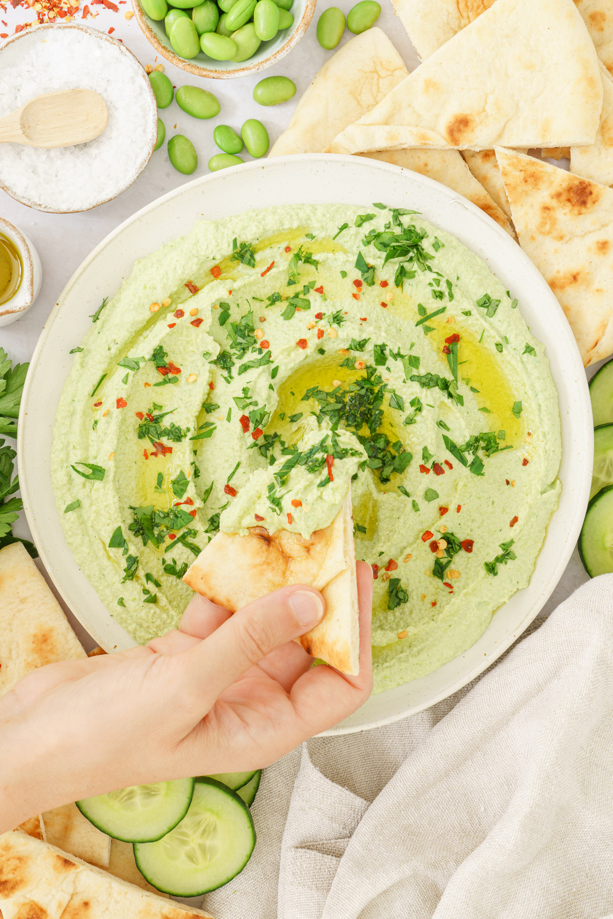 top view image of a hand tossing pita bread in to an edamame hummus