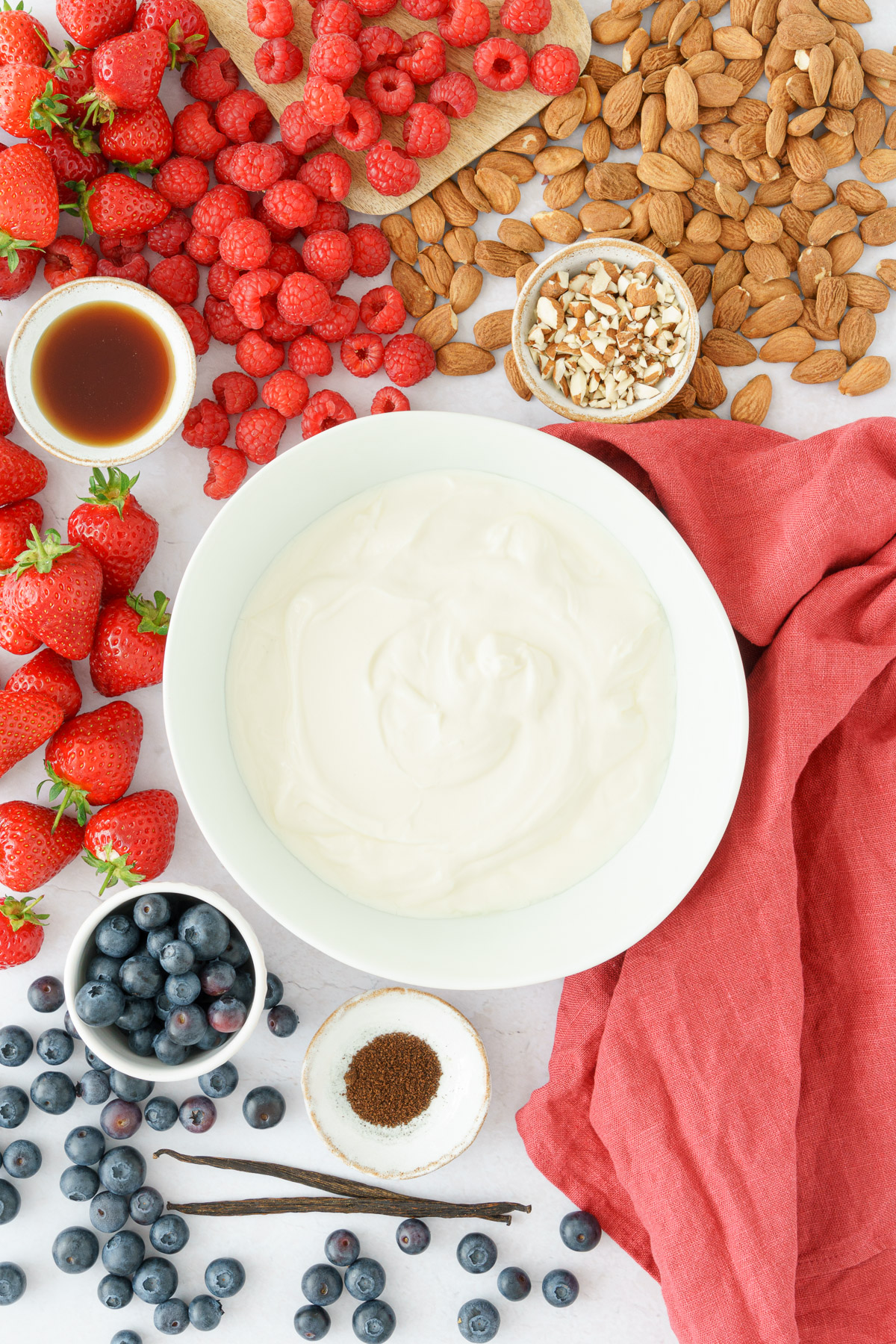 top view image of frozen yogurt bark ingredients yogurt on a round plate, with strawberries, raspberries, blueberries, nuts and maple syrup on a table