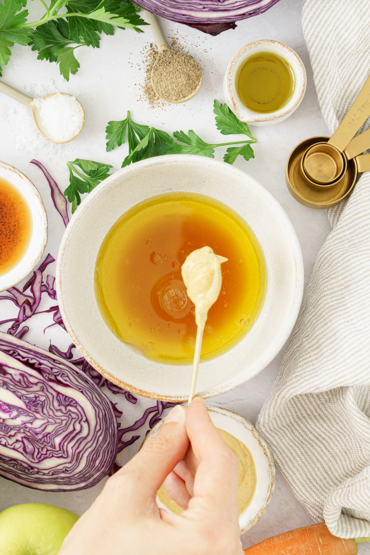 an image of a hand adding a dijon  mustard to the mixture of olive oil, apple cider vinegar, maple syrup and salt
