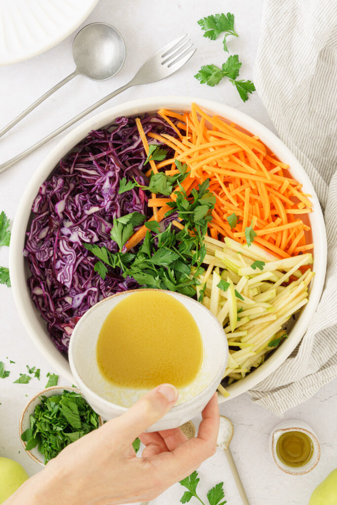closed up image of bowl with combined thinly sliced red cabbage, apples, and carrots. A hand pouring the dressing