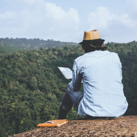man reading a book in nature with green mountains ahead of him