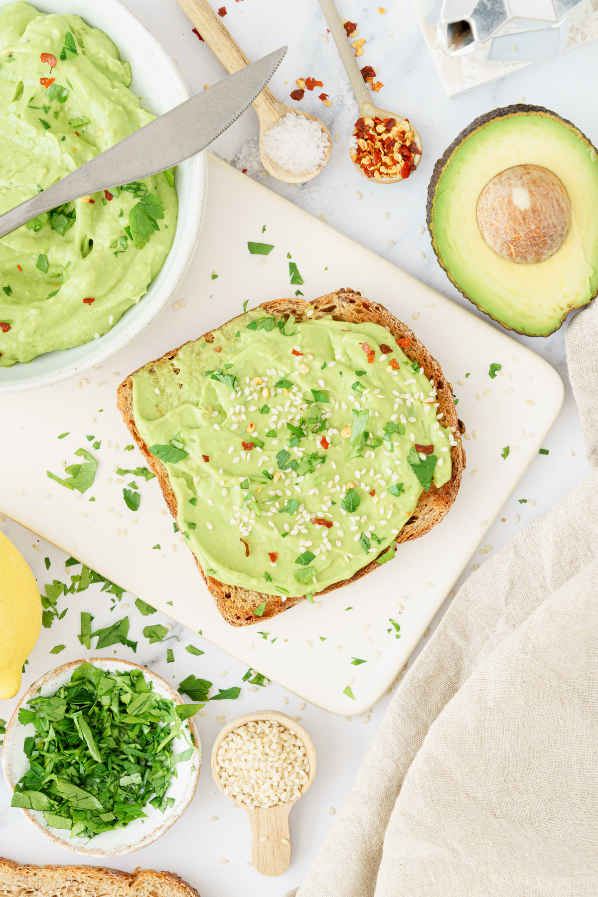 Single slice of bread on a cutting board, generously spread with avocado spread and garnished with sesame seeds, fresh herbs, and chili flakes.