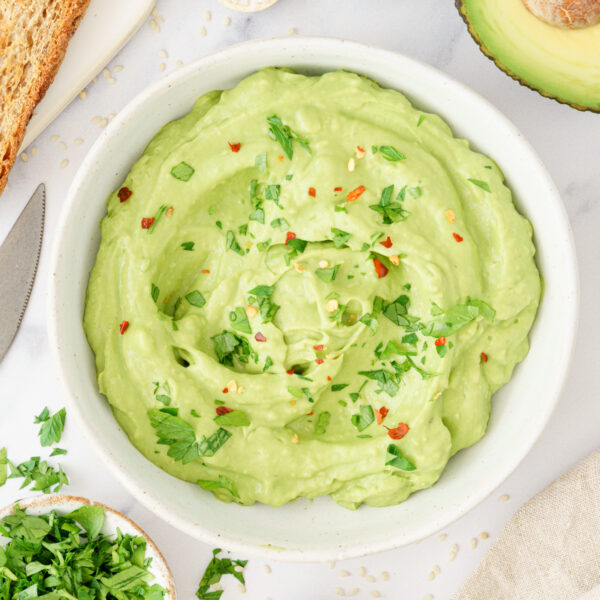 Bowl filled with creamy avocado spread, garnished with red chili flakes and fresh herbs.