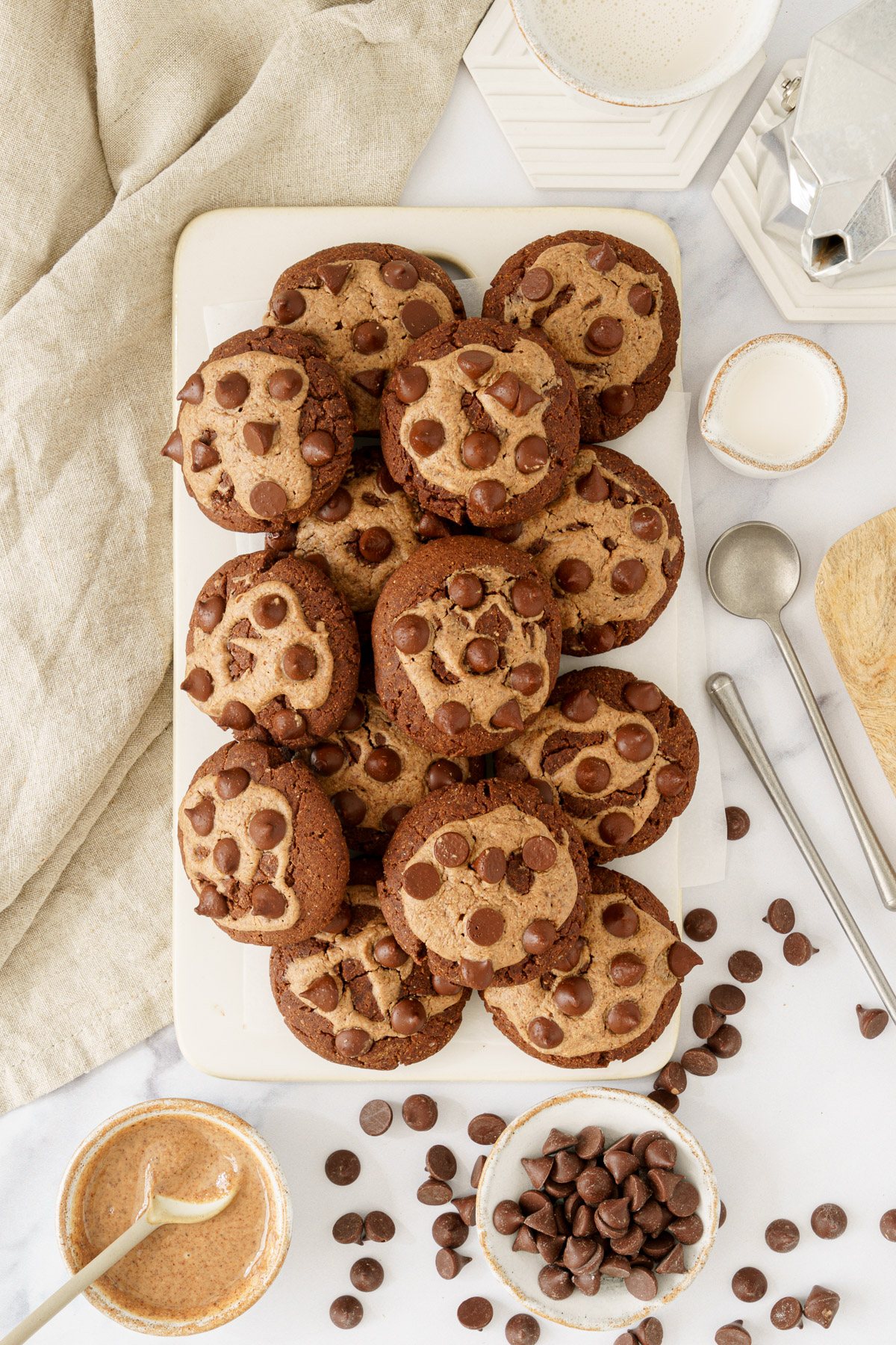 top down view image of a cutting board with a batch of freshly baked chocolate chickpea flour cookies, featuring chocolate chips and a mixture of delicious toppings nearby