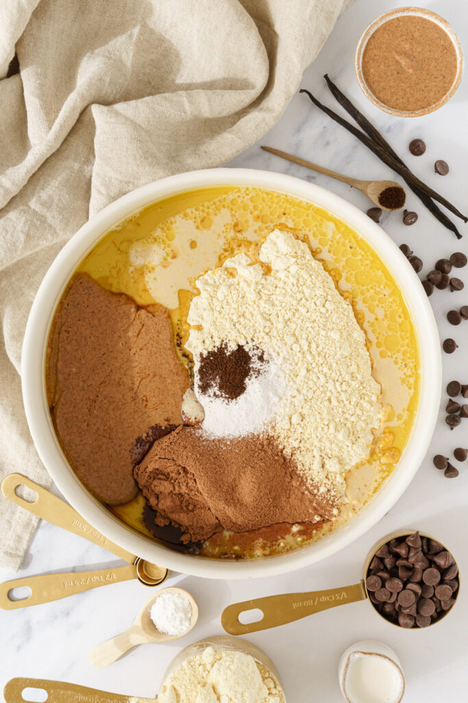 top-down view of a bowl filled with the ingredients for chocolate chickpea cookies: chickpea flour, cocoa powder, virgin oil, with a cup of chocolate chips positioned nearby