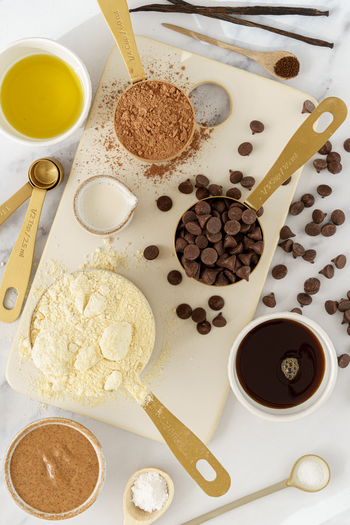 top-down view of a cutting board with a variety of ingredients arranged neatly. The ingredients include chocolate chips, chickpeas, sea salt, vanilla powder, baking powder, virgin oil, maple syrup, and almond milk