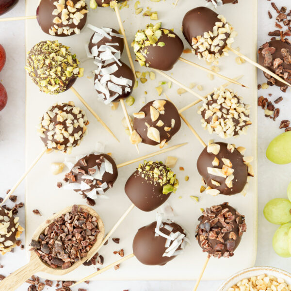 cropped image of a top-down view of chopping board displaying skewers of chocolate-covered grapes, garnished with nuts, shredded coconut, and crushed chocolate