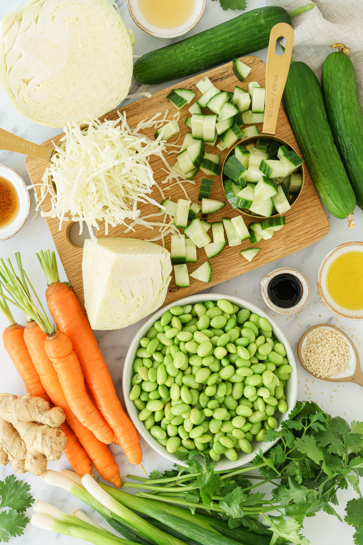 top-down view image displaying ingredients for an edamame salad, including edamame beans, carrots, ginger, diced cucumber, green onions, sesame seeds, shredded cabbage, and cilantro, soy sauce, and sesame oil