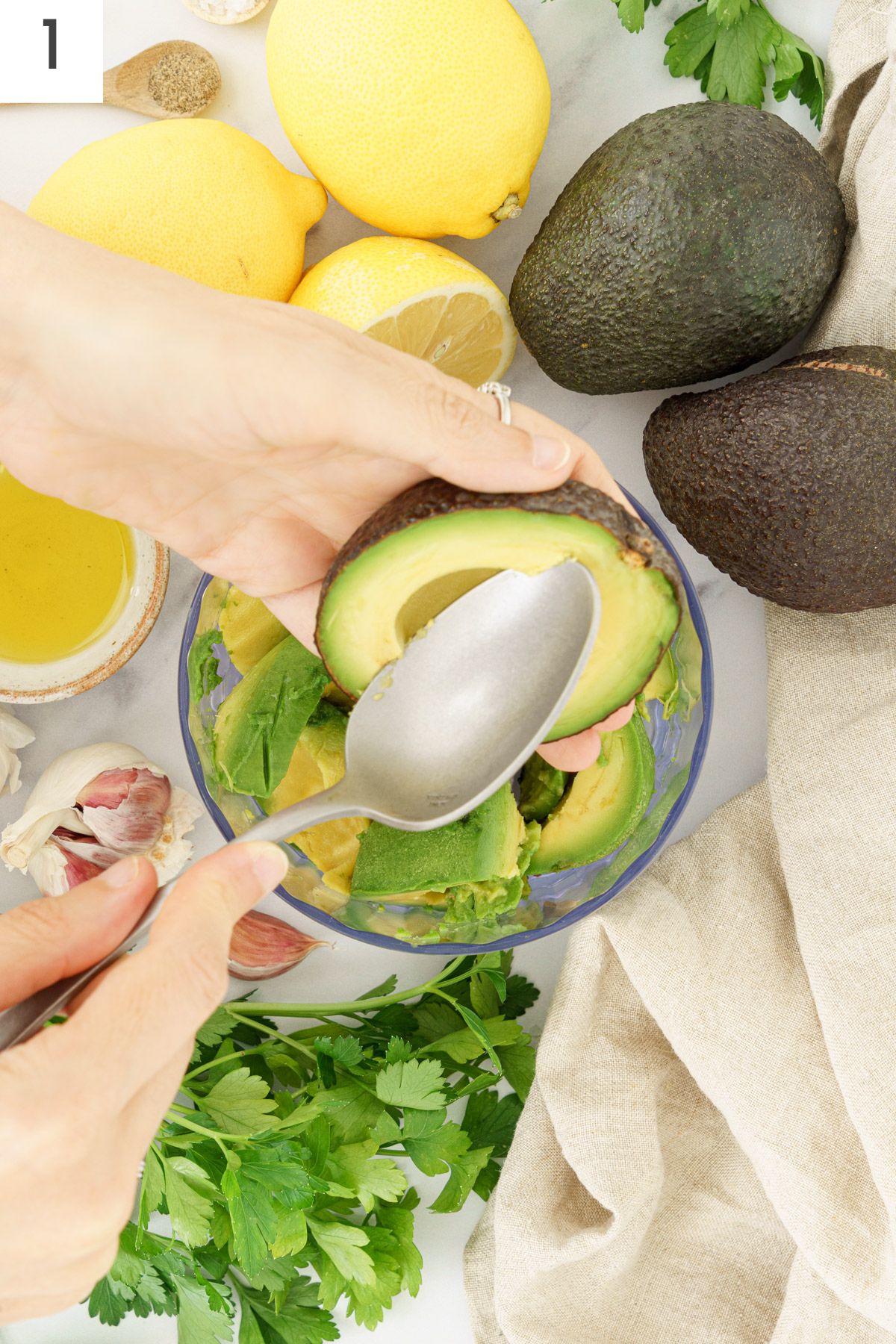Hands scooping out avocado in a food processor with a spoon.