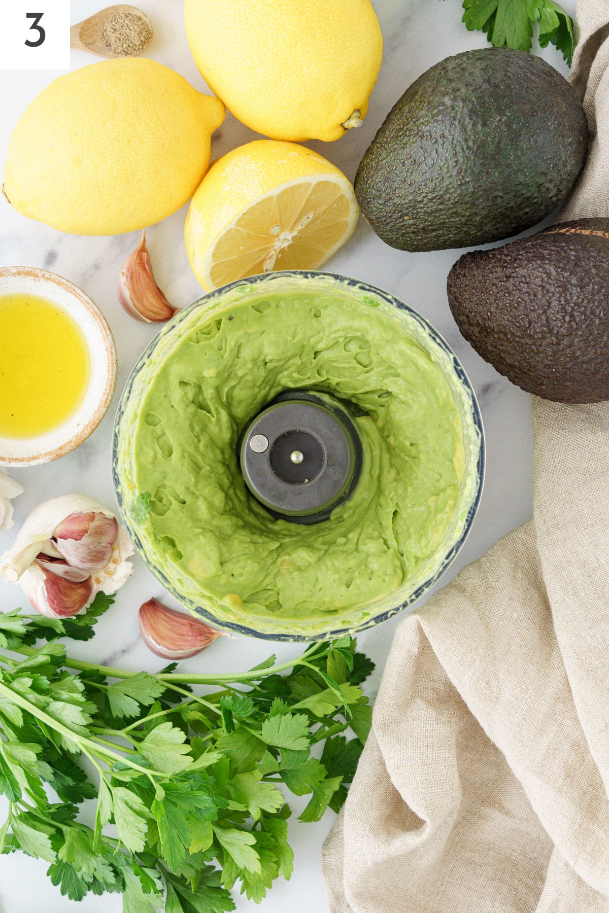 Food processor containing pulsed avocados, surrounded by whole avocados, lemons, fresh herbs, and garlic.