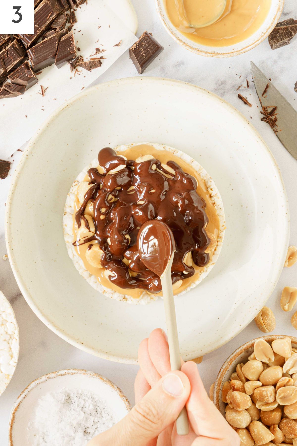Hand spreading melted chocolate on top of the peanuts and peanut butter.
