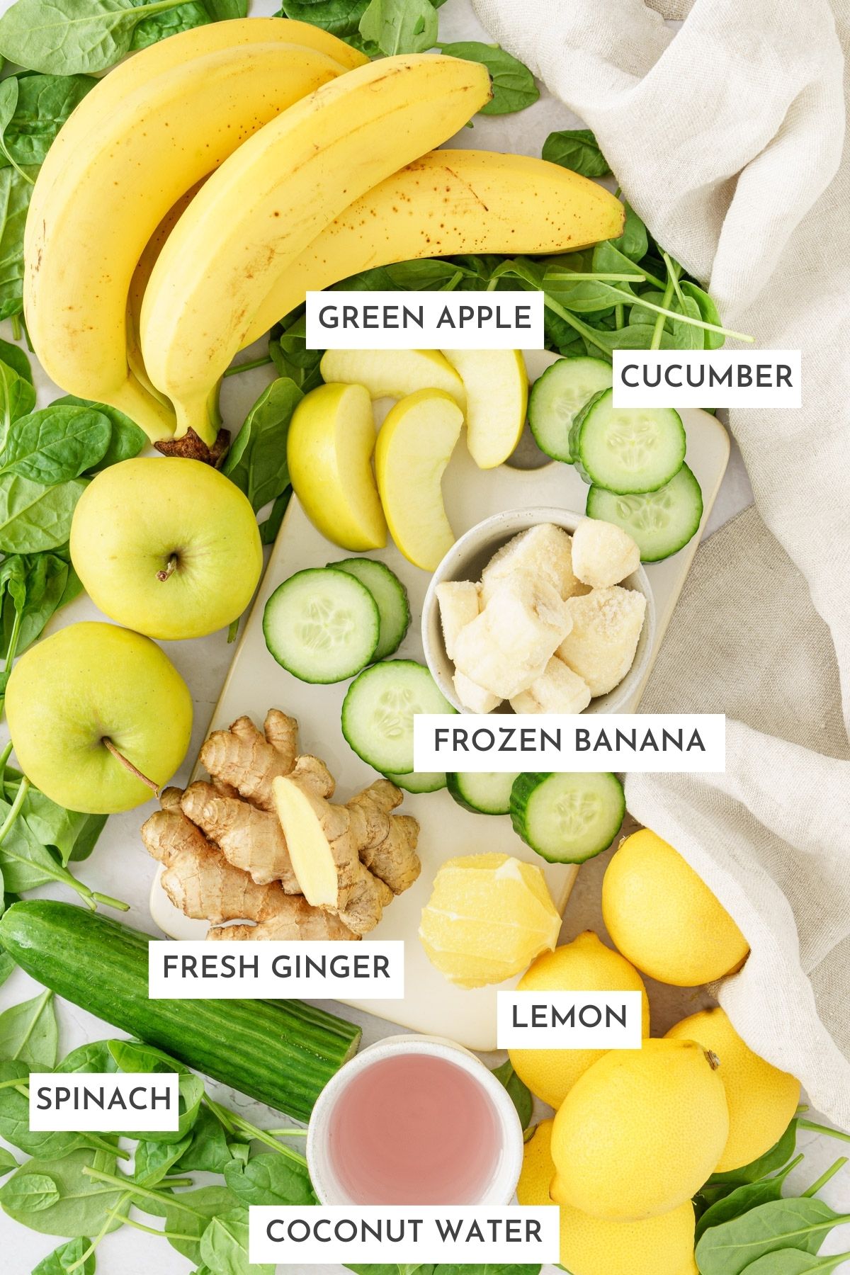 Ingredients laid on a table: green apple, cucumber, spinach, fresh ginger, lemon, coconut water.