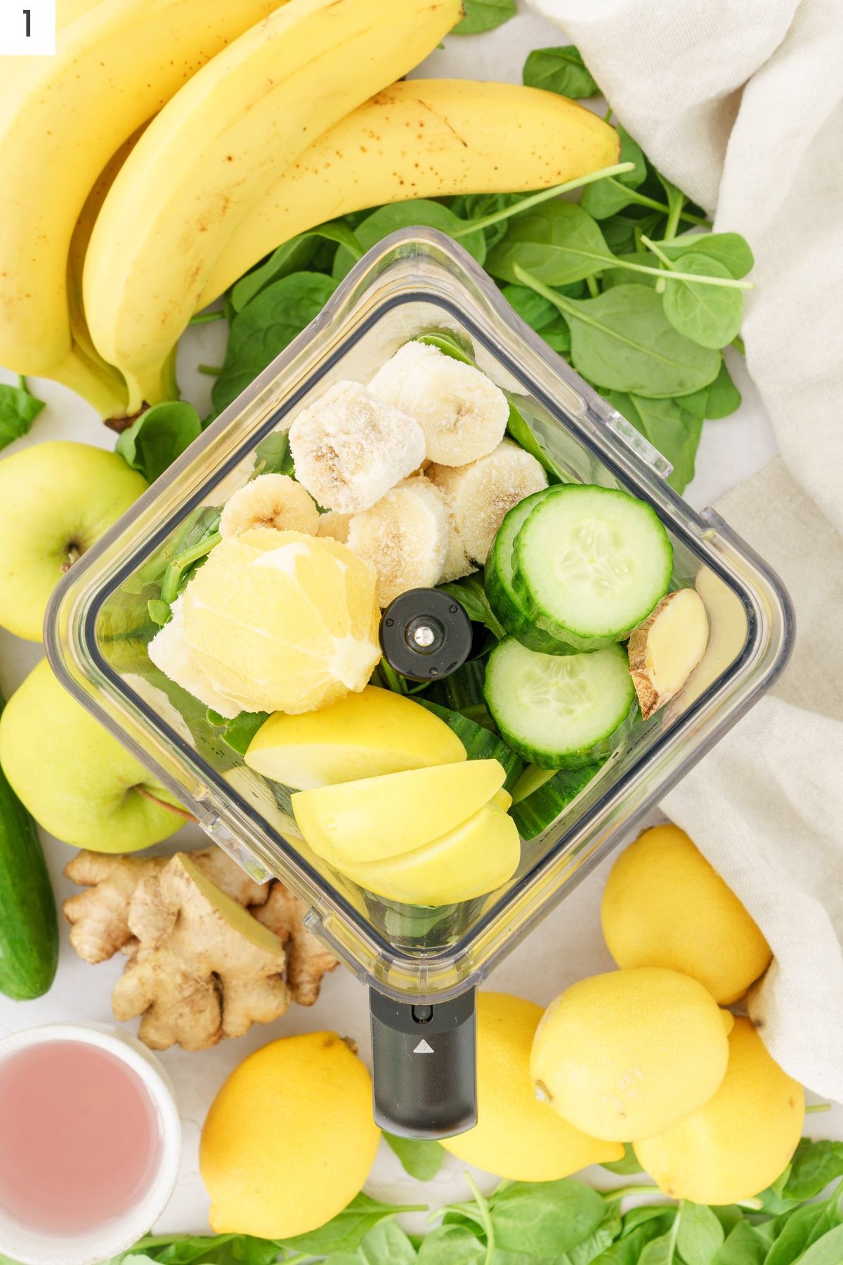 Blender with frozen banana, sliced cucumber, peeled lemon, cut green apple sliced ginger, and spinach.