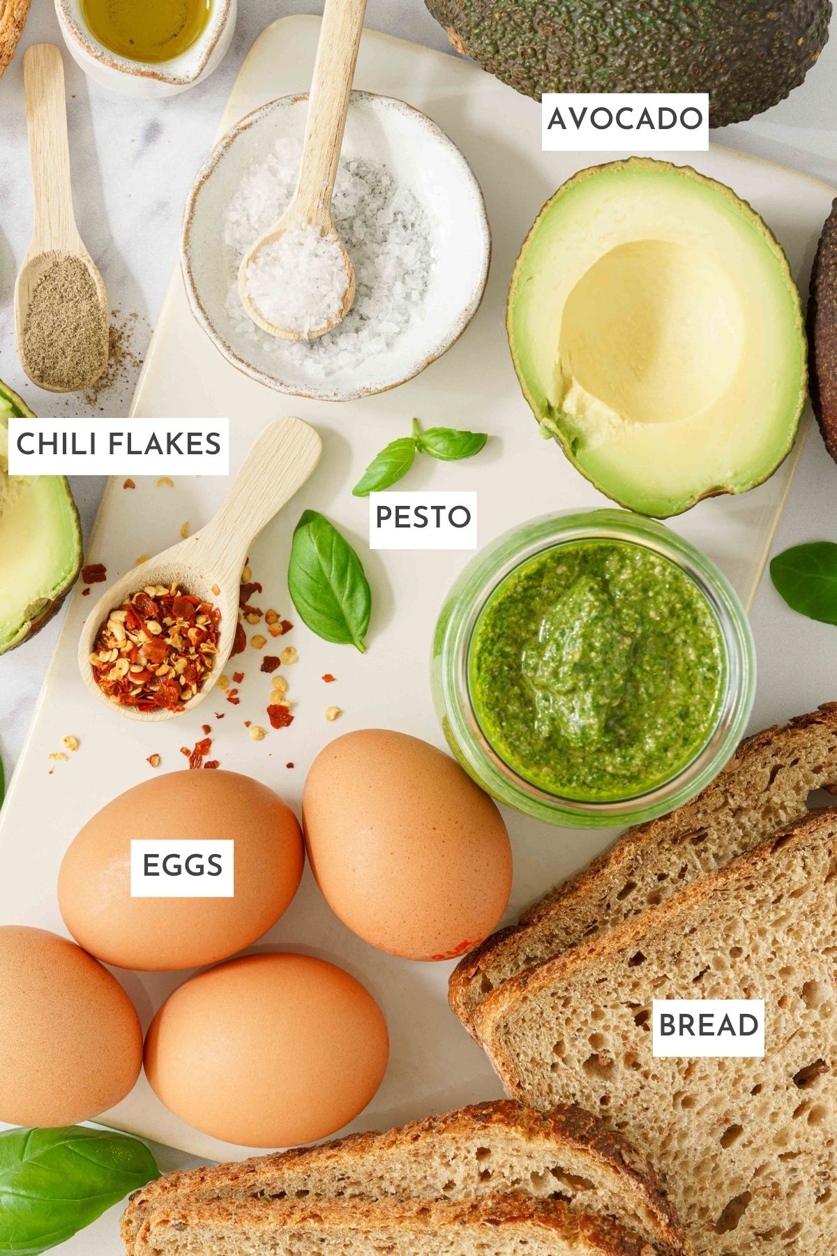 All ingredients laid out on a table: avocado, chili flakes, pesto, eggs, bread.