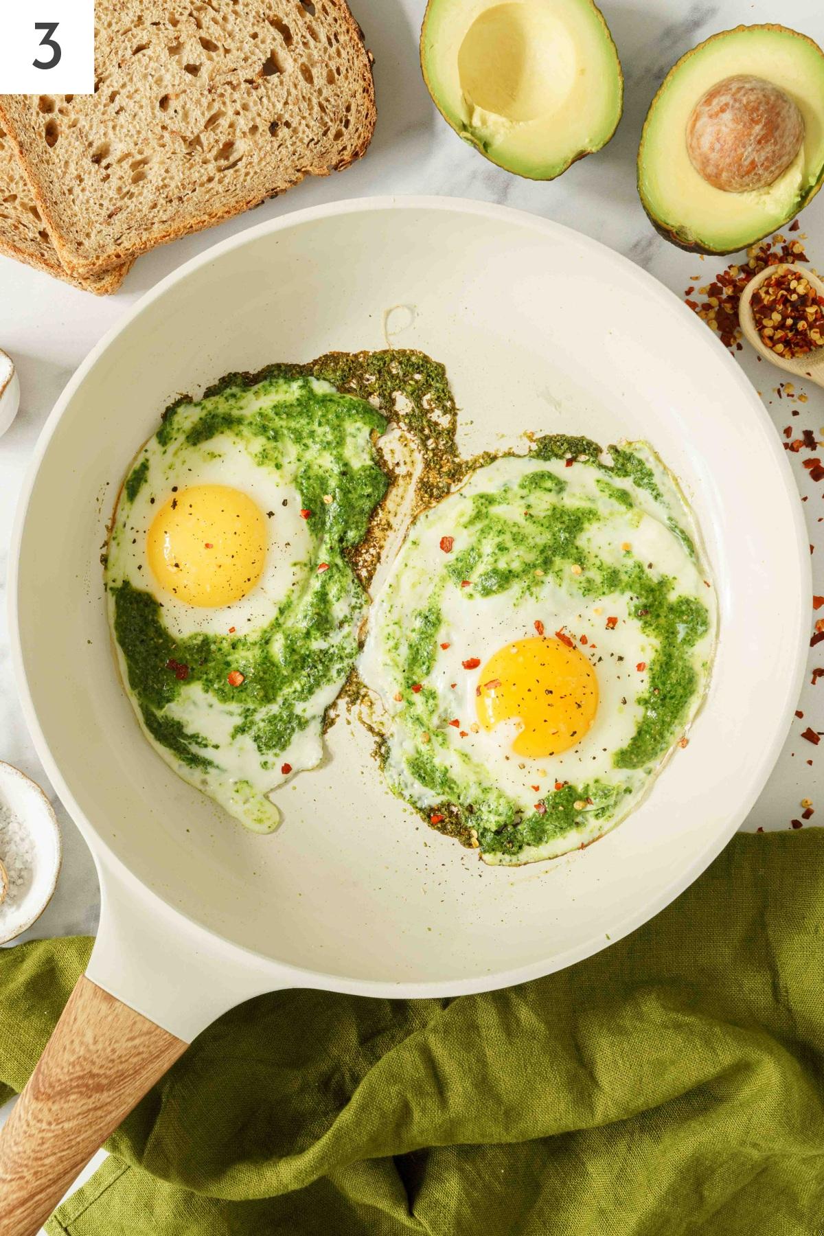 A pan with spreaded pesto and eggs sprinkled with chili flakes.