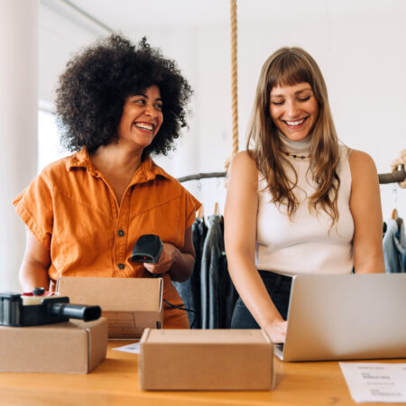 Two women working together and laughing in front of a laptop and boxes ready to be shipped.