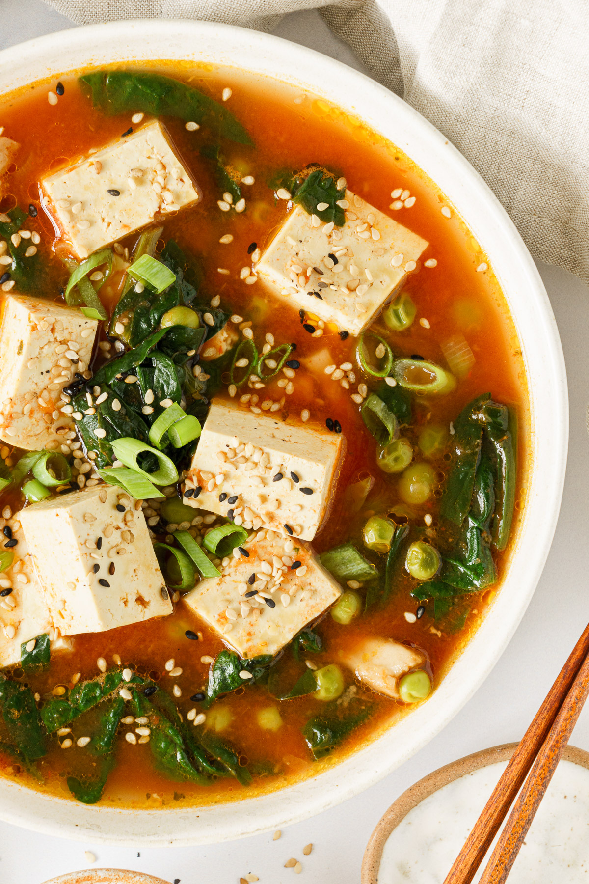A bowl of tofu soup sprinkled with green onions, sesame seeds, and chili flakes.