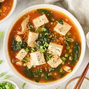 A bowl of tofu soup garnized with chopped green onions, sesame seeds, and chili flakes.