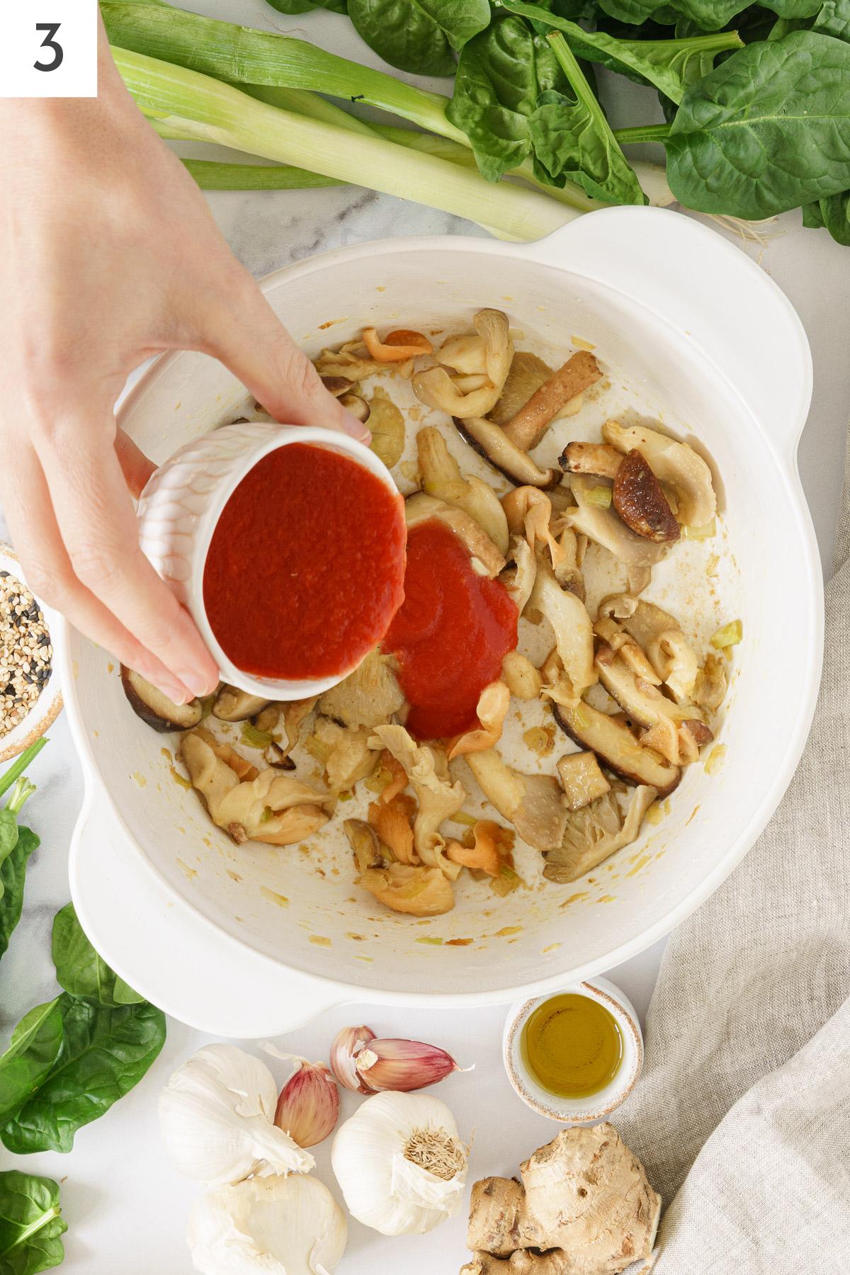 A hand pouring tomato sauce into the sauteed ingredients.