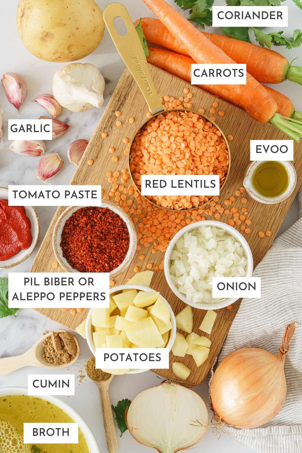 Laid out ingredients of turkish lentil soup: coriander, carrots, garlic, evoo, red lentils, tomato paste, aleppo peppers, onion, potatoes, cumin, and broth.