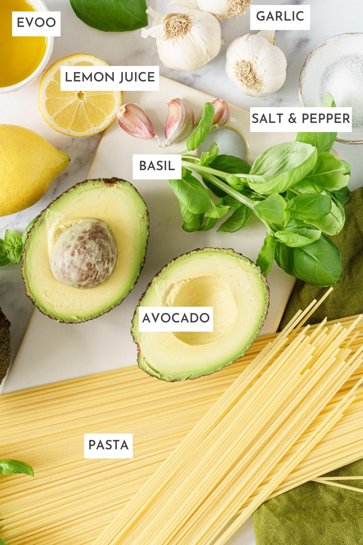 Laid out ingredients of avocado pasta sauce: Evoo, garlic, lemon juice, salt and pepper, basil, avocado, and pasta.