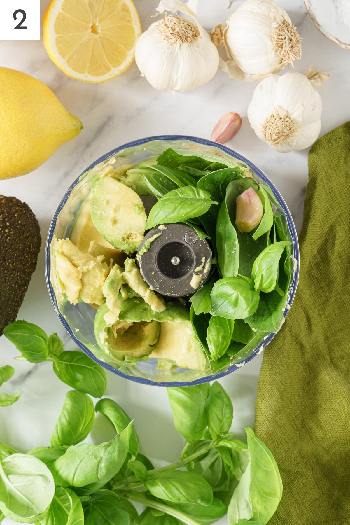 Food processor containing with avocado, garlic, lemon juice, olive oil, pasta water, and basil leaves