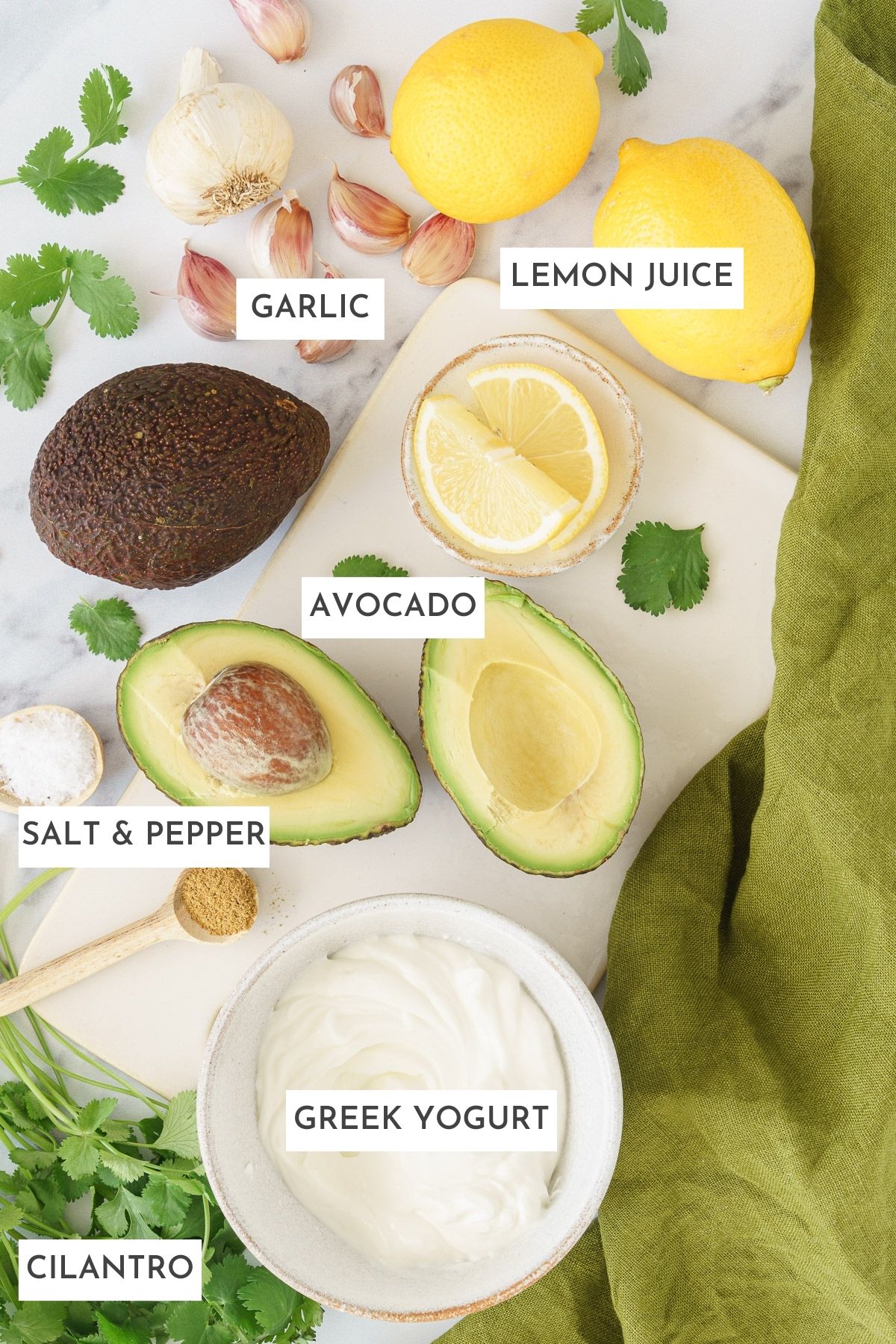 Laid out ingredients of avocado sauce: Garlic, lemon juice, avocado, salt and pepper, greek yogurt, and cilantro.