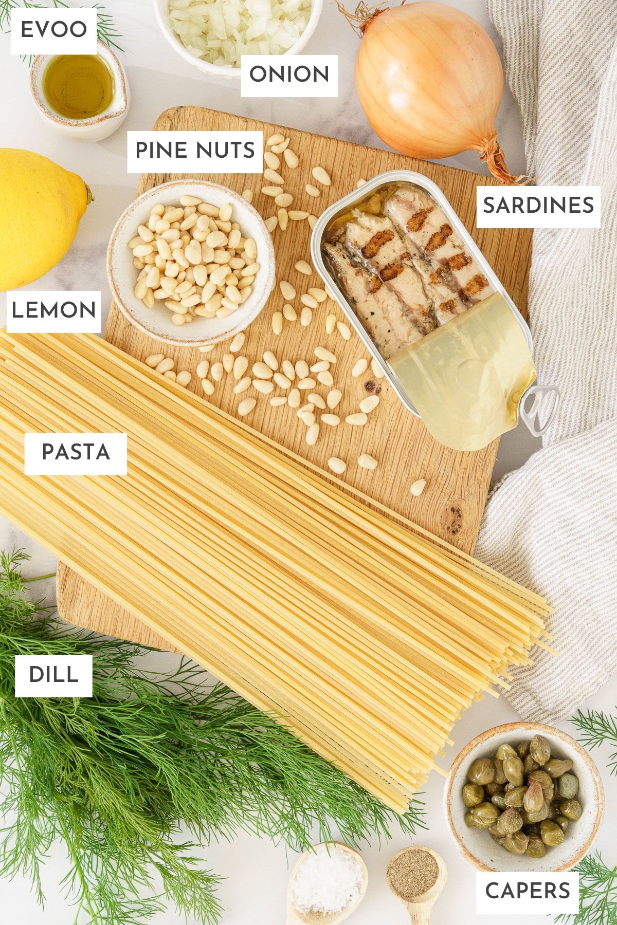 Laid out ingredients of mediterranean sardine pasta; evoo, onion, pine nuts, sardines, lemon, pasta, dill, and capers.